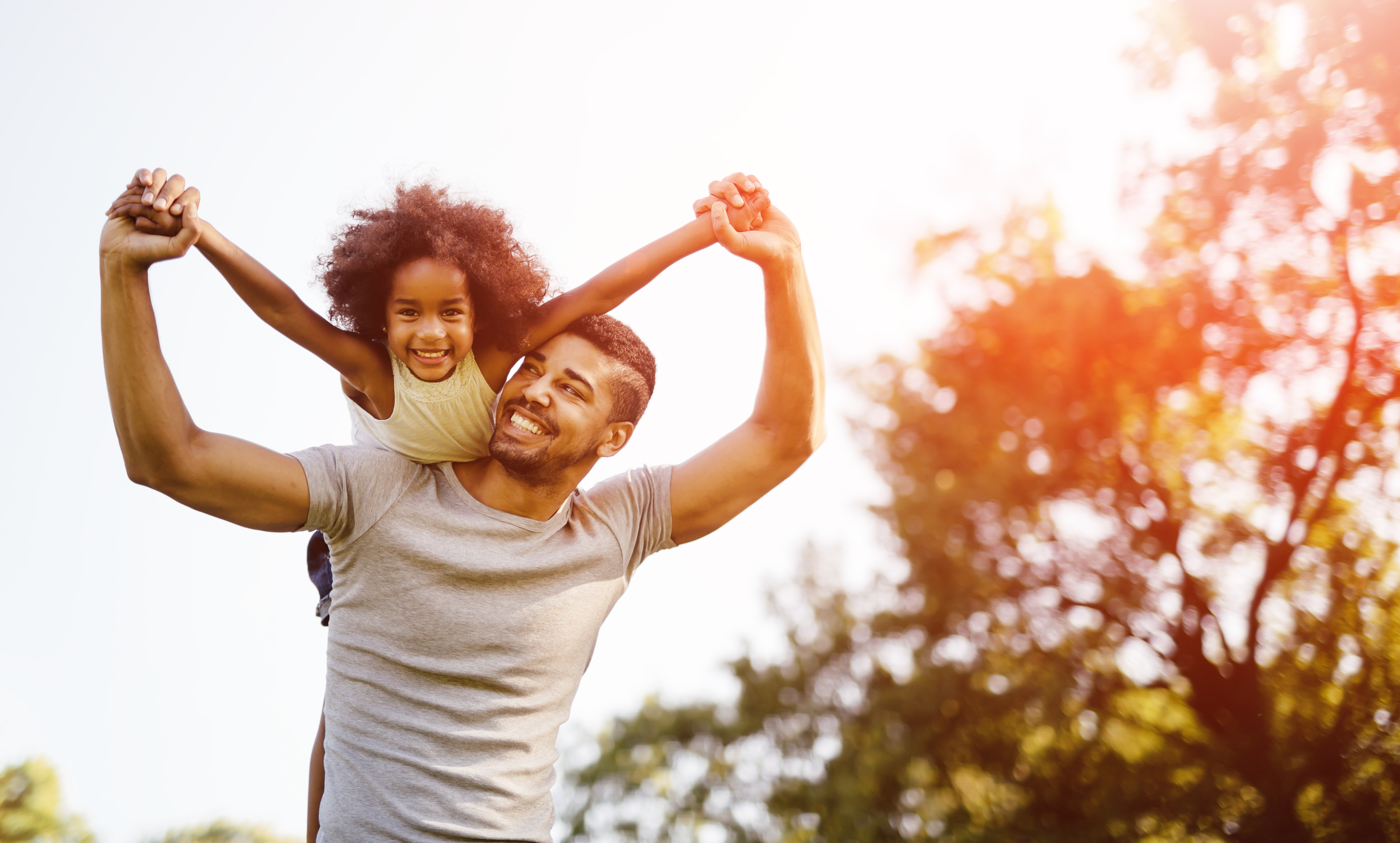 A father lifts his child up on his shoulders with her arms outstretched like an airplane